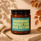 Amber jar candle with teal label reading "STATUS: BURNING BRIGHT FOR OUR FUTURE".
