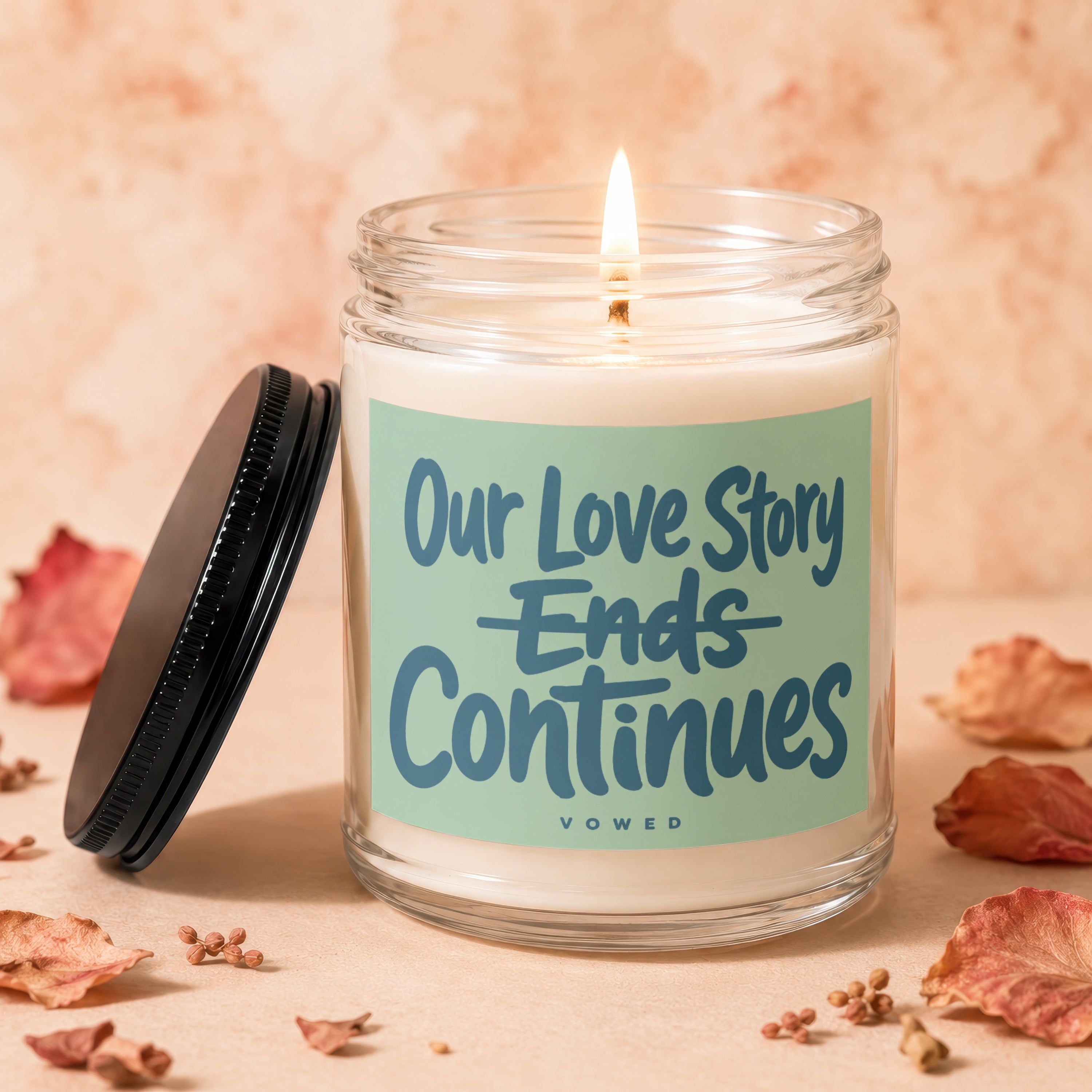Glass candle with burning wick and a label reading "Our Love Story Ends Continues VOWED".