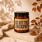Brown glass candle labeled "Smells Like HE HIT THE JACKPOT" on a stone block.