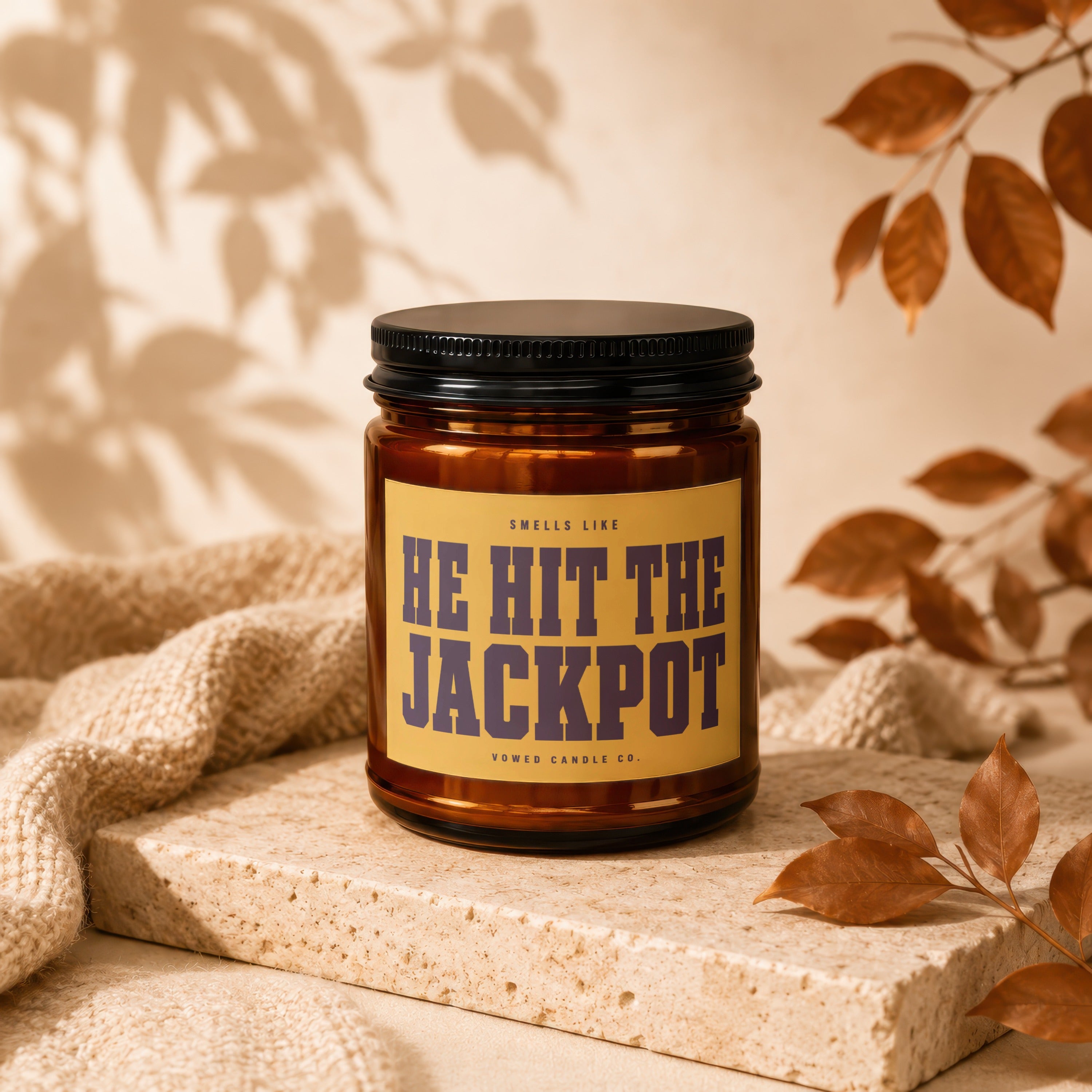 Brown glass candle labeled "Smells Like HE HIT THE JACKPOT" on a stone block.