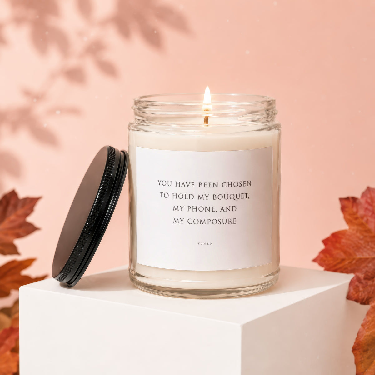 Glass candle with lit wick and label reading "You have been chosen to hold my bouquet, my phone, and my composure."