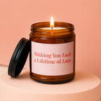 Amber jar candle with lit wick and pink label reading "Wishing You Luck a Lifetime of Love."