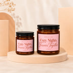 Two glass candles with pink labels reading "Cozy Nights, Forever Together".
