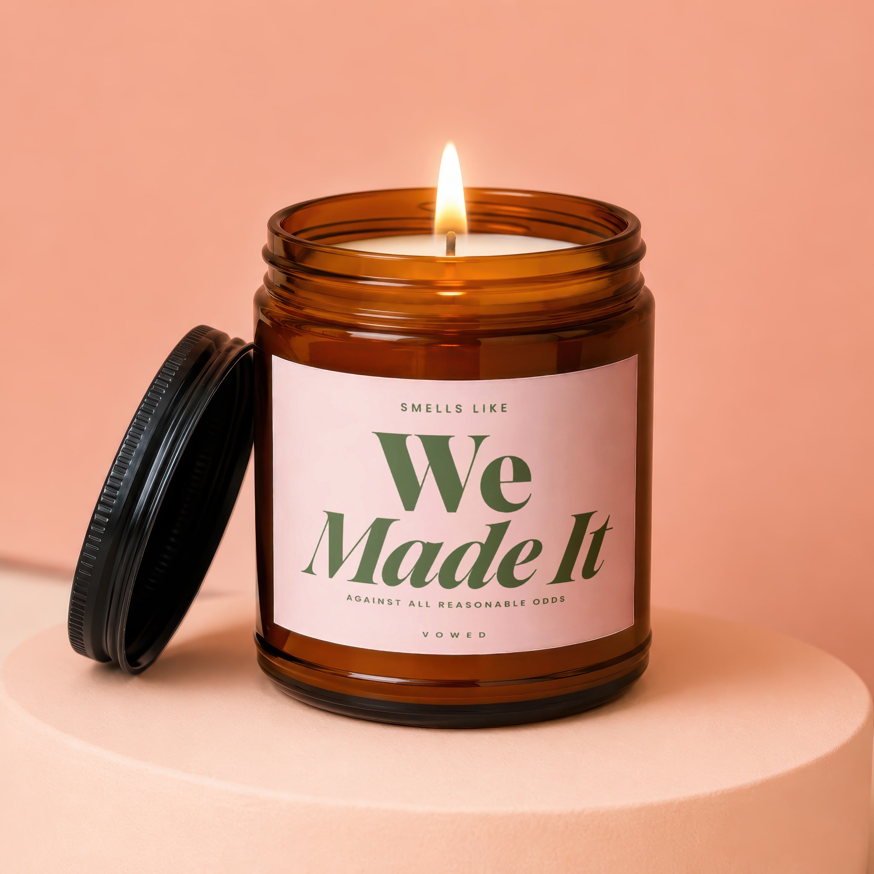 Amber jar candle with lit wick and pink label reading "Smells Like We Made It".