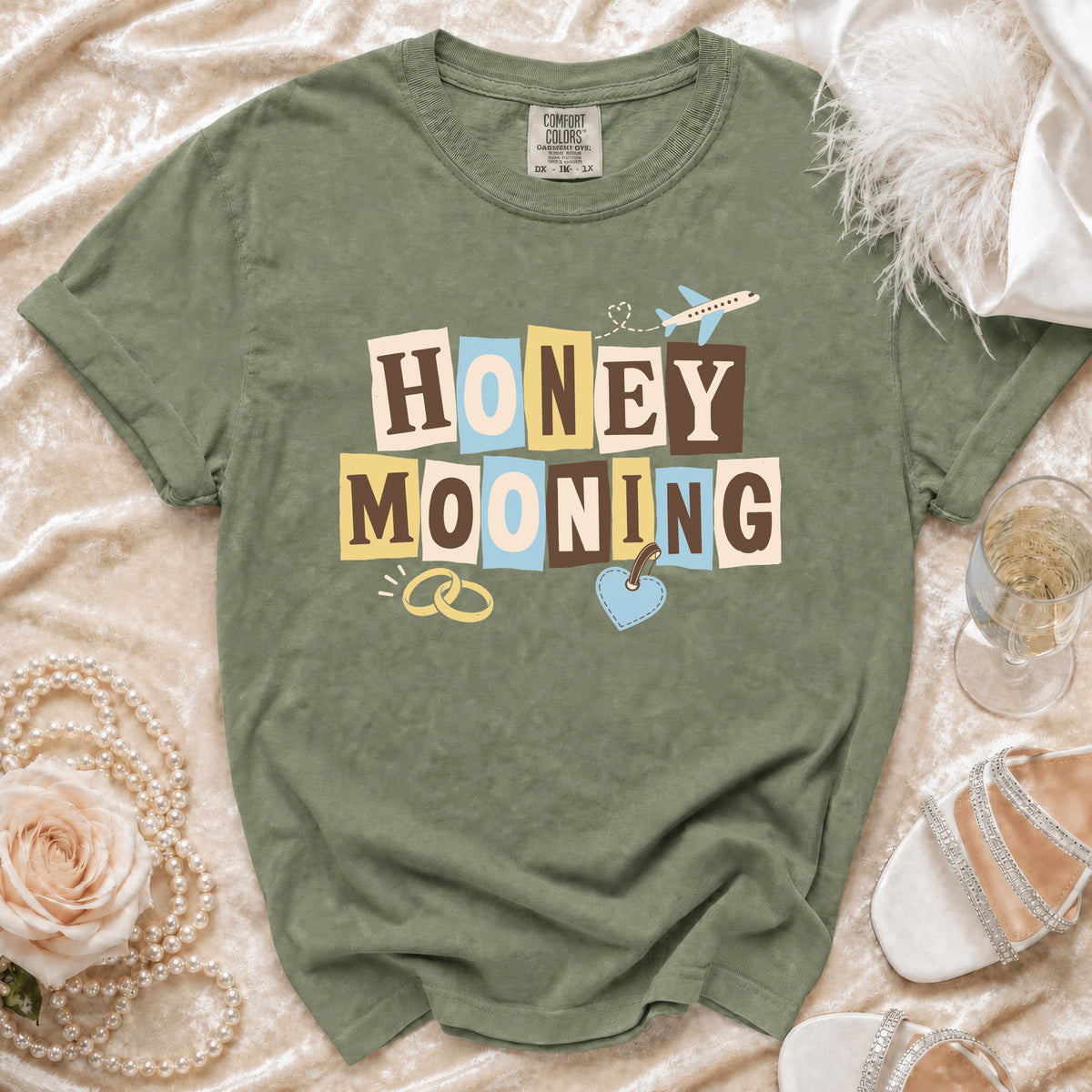 Olive green t-shirt with "HONEY MOONING" text, wedding rings, and airplane graphic.