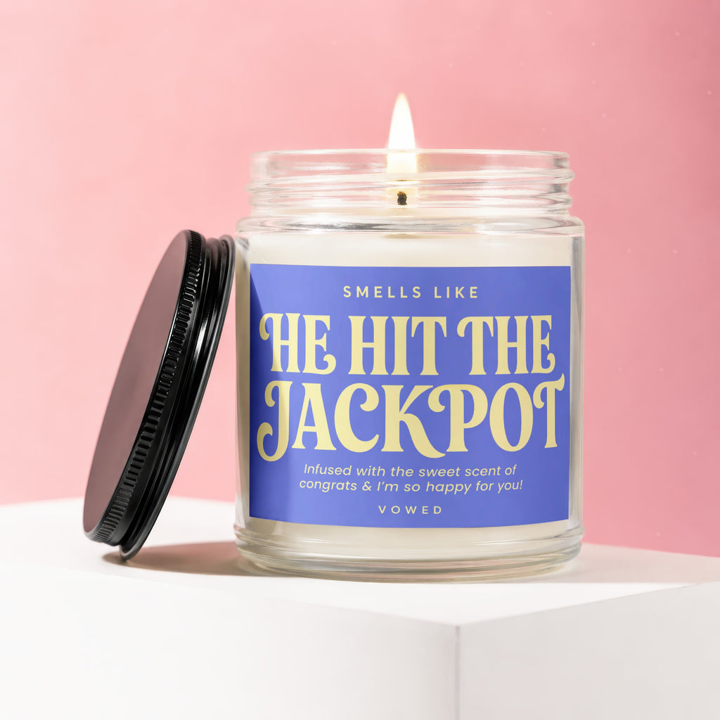 Glass jar candle with a lit wick and purple label reading "He Hit The Jackpot"