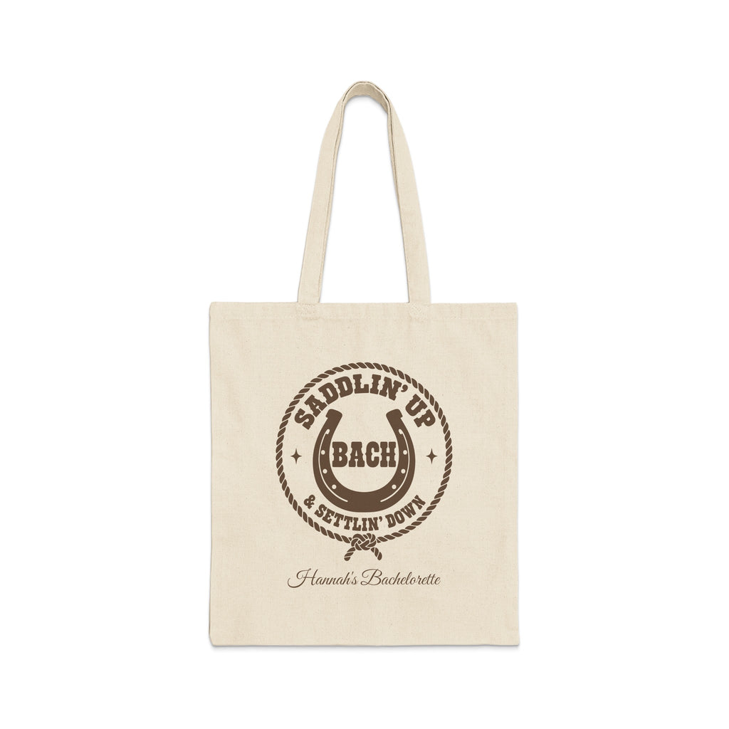 Off-white canvas tote bag with brown rodeo-themed text, horseshoe, and rope graphic on the front.
