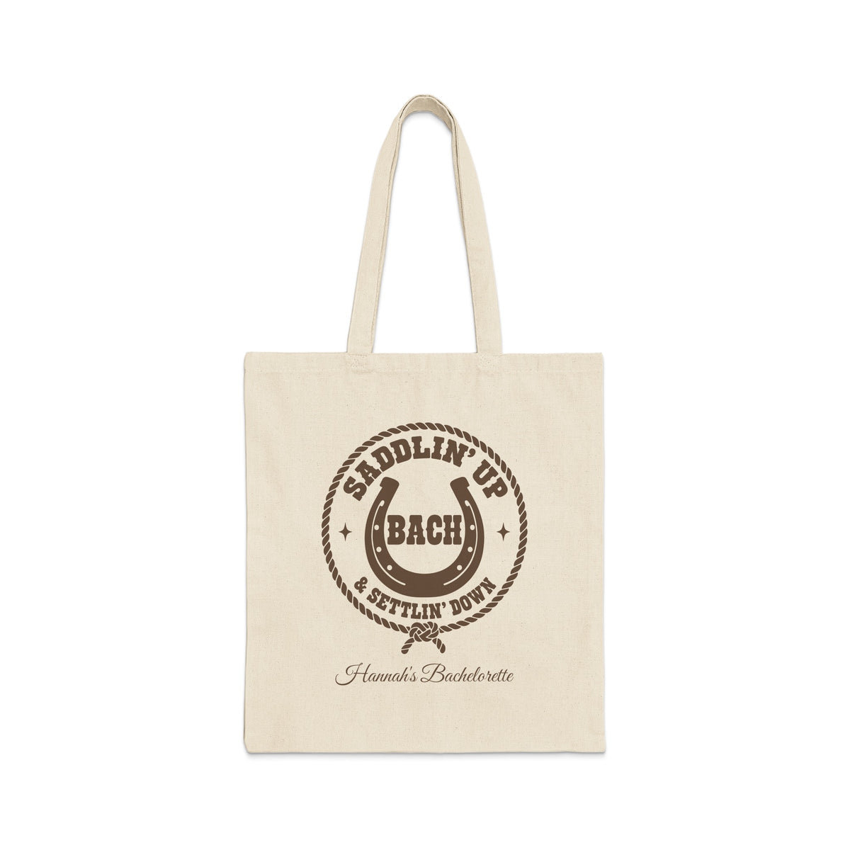 Off-white canvas tote bag with brown rodeo-themed text, horseshoe, and rope graphic on the front.