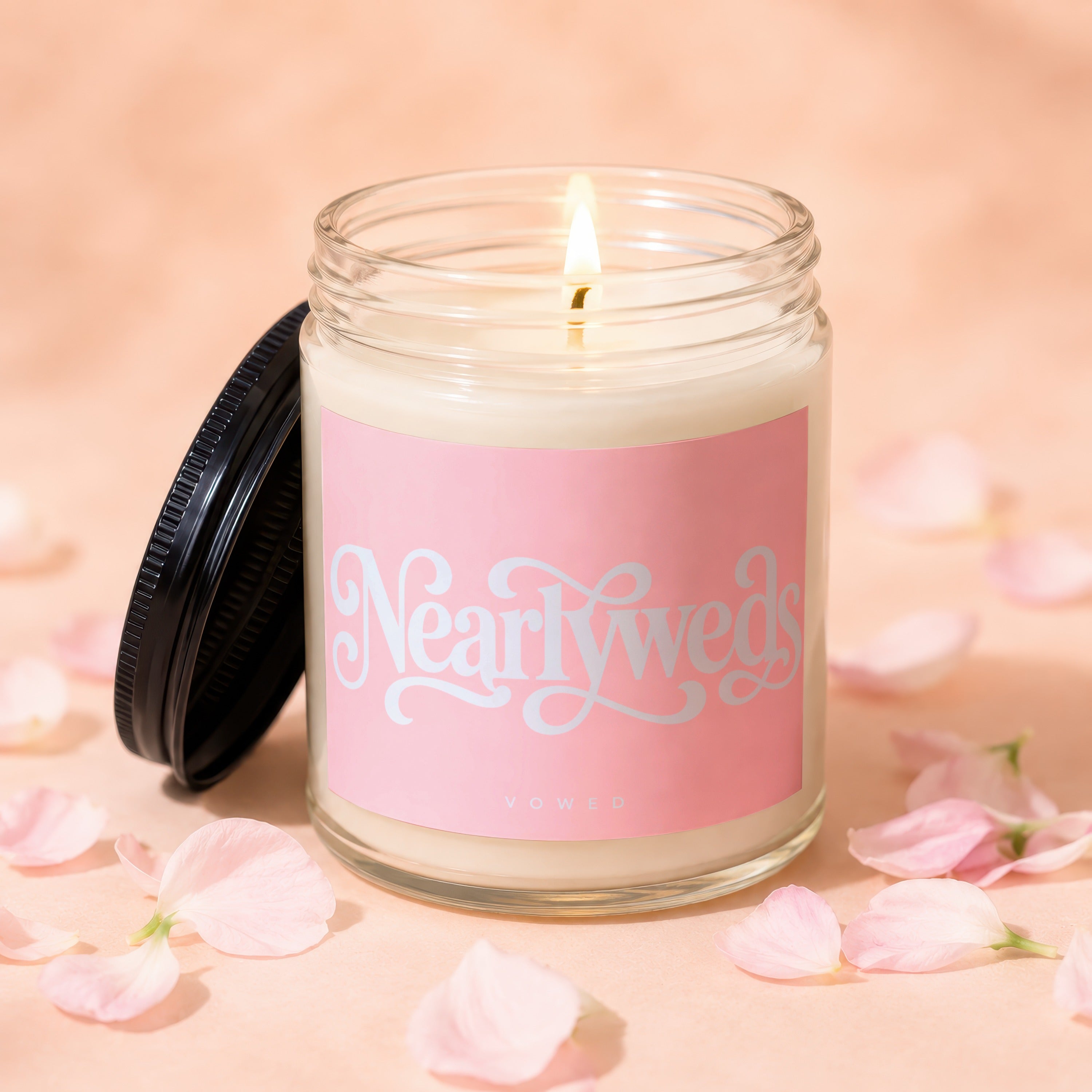 Lit candle in a glass jar with a pink label reading "Nearlyweds VOWED", surrounded by petals.