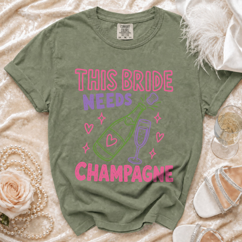 Olive green t-shirt with "This bride needs champagne" graphic in pink and purple.
