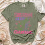 Olive green t-shirt with "This bride needs champagne" graphic in pink and purple.