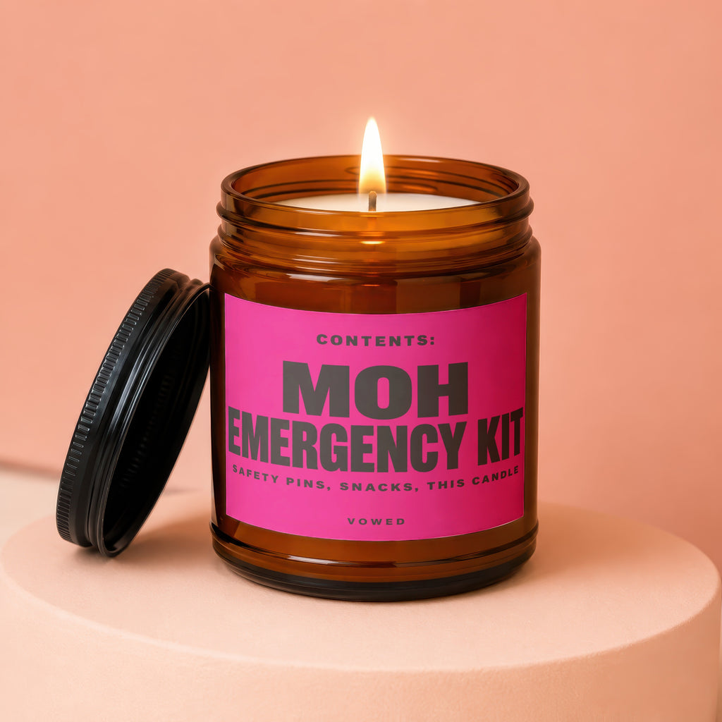 Amber jar candle with lit wick and pink label reading: MOH Emergency Kit, Safety Pins, Snacks, This Candle
