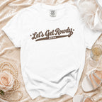 A white crewneck t-shirt with brown script text reading "Let's Get Rowdy BRIDE" rests on a textured cream surface with accessories.
