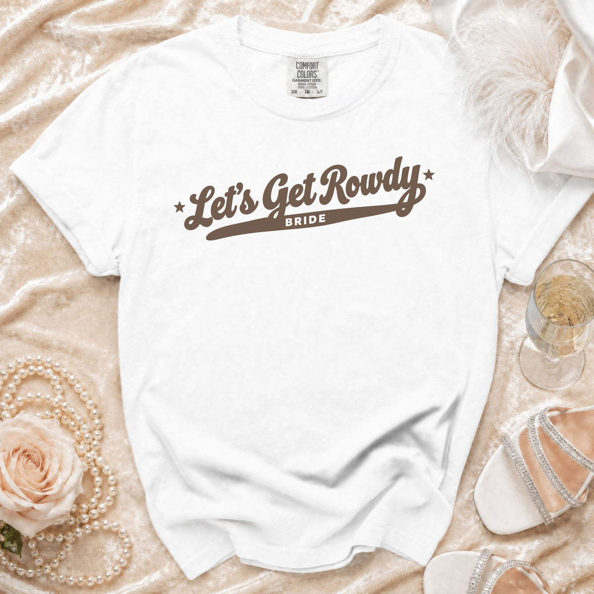 A white crewneck t-shirt with brown script text reading "Let's Get Rowdy BRIDE" rests on a textured cream surface with accessories.