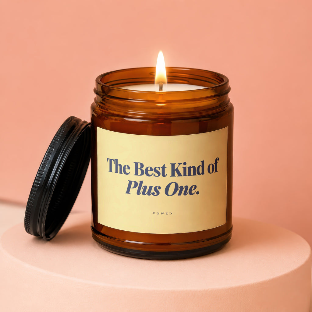 Amber jar candle with lit flame and lid. Label reads "The Best Kind of Plus One."