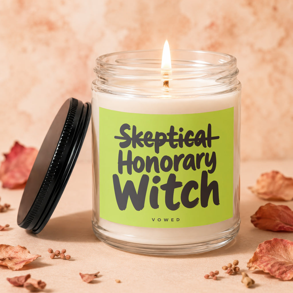 Skeptical Honorary Witch candle with lit wick and black lid, surrounded by dried leaves.