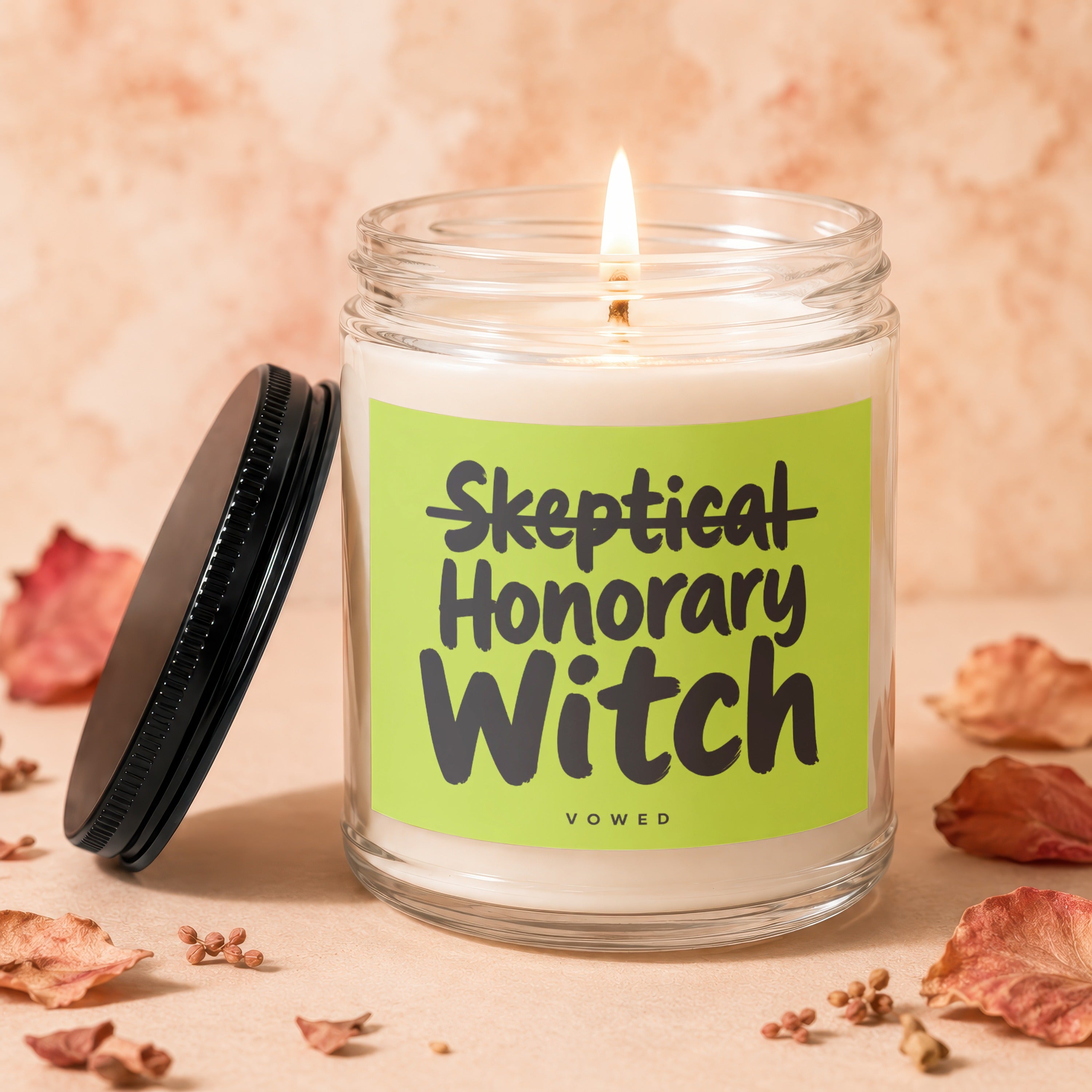 Skeptical Honorary Witch candle with lit wick and black lid, surrounded by dried leaves.