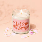 Candle with "Here come the grooms" text and "VOWED" on pink label, surrounded by petals.
