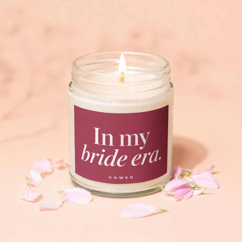 Lit candle with maroon label reading "In my bride era." surrounded by petals.