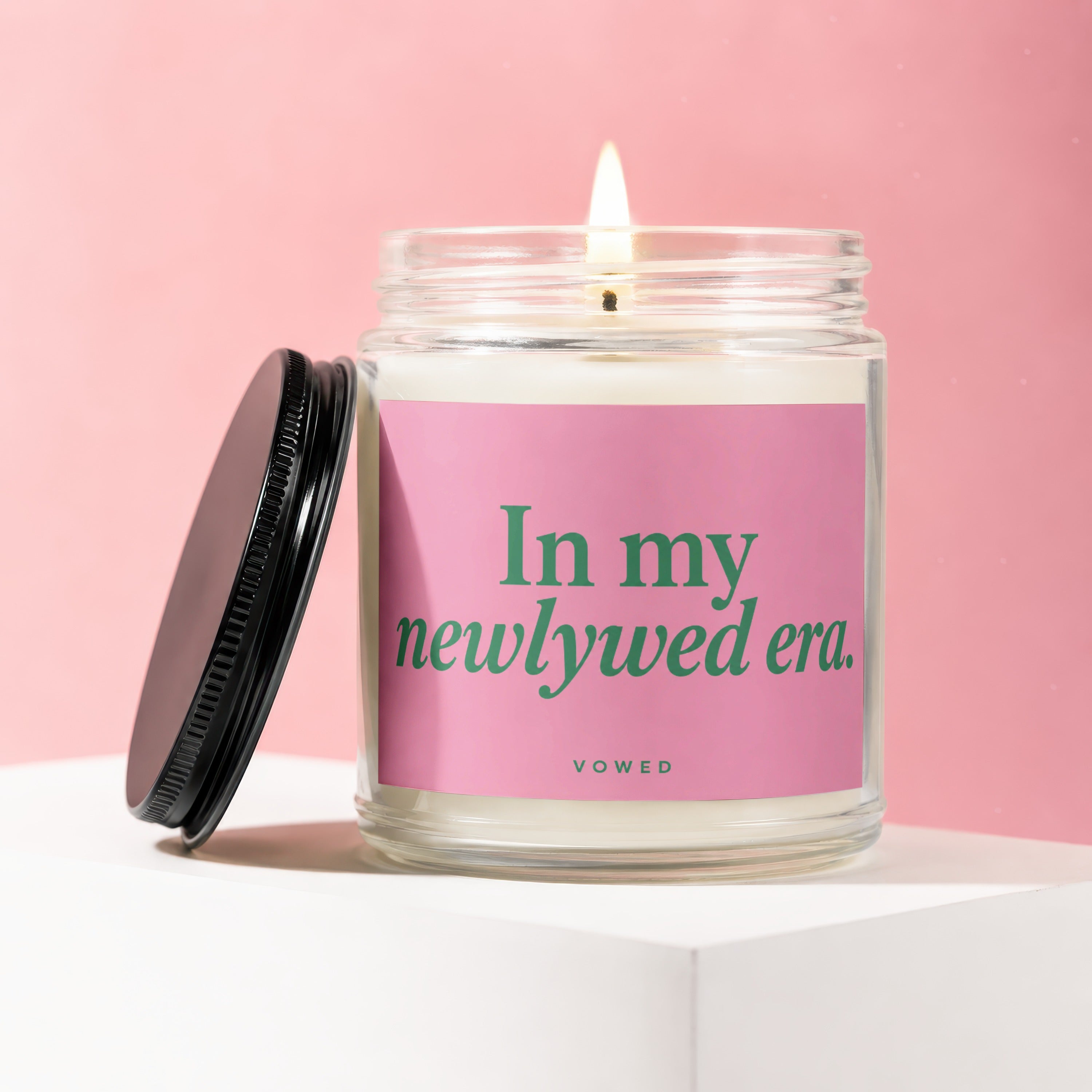 Lit candle in a glass jar with "In my newlyweds era. vowed" on a pink label with a black lid.