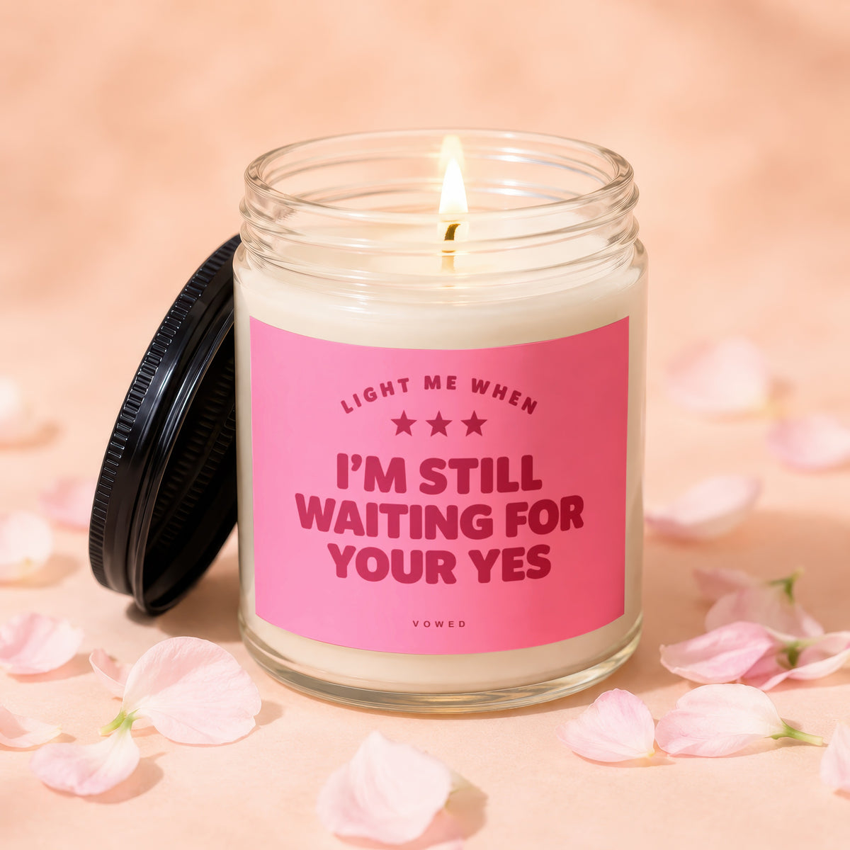 Glass candle with lit wick and pink label that reads "Light me when I'm still waiting for your yes".