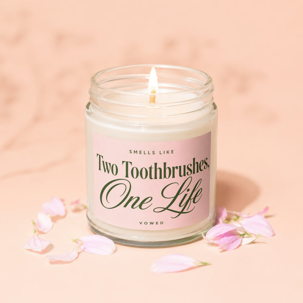 Lidded candle with pink label reading "Smells like Two Toothbrushes, One Life" surrounded by petals.