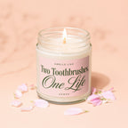 Lidded candle with pink label reading "Smells like Two Toothbrushes, One Life" surrounded by petals.
