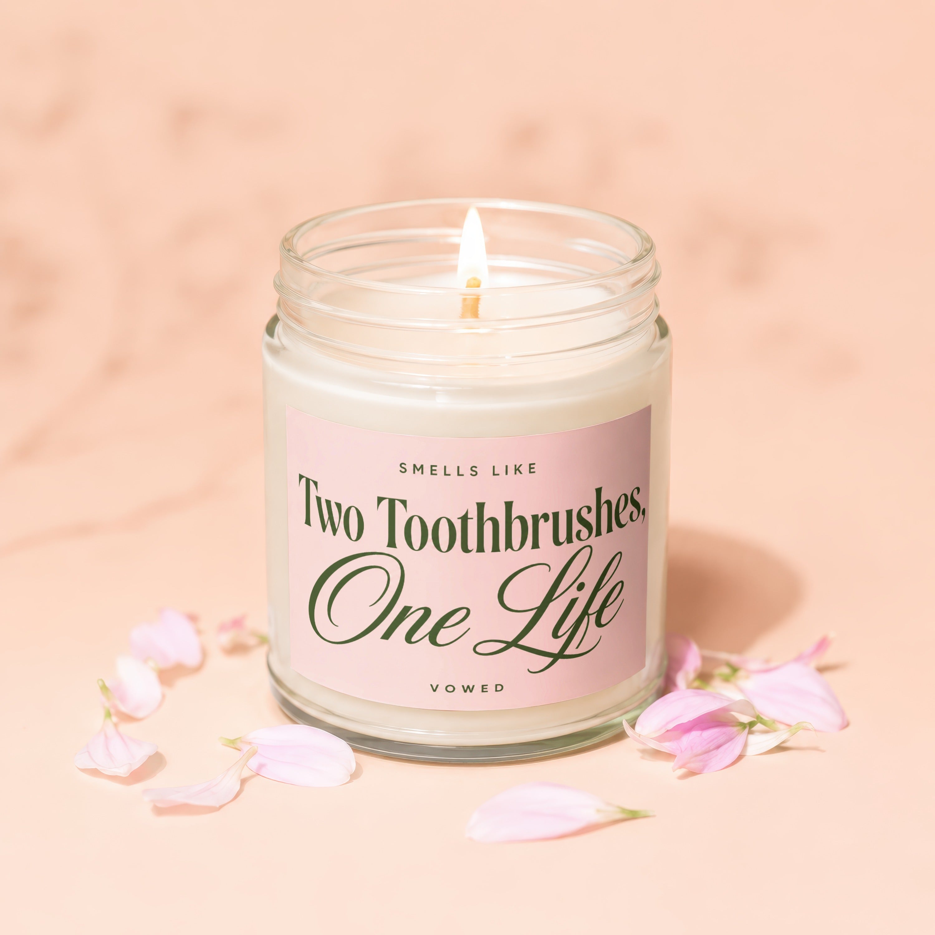 Lidded candle with pink label reading "Smells like Two Toothbrushes, One Life" surrounded by petals.