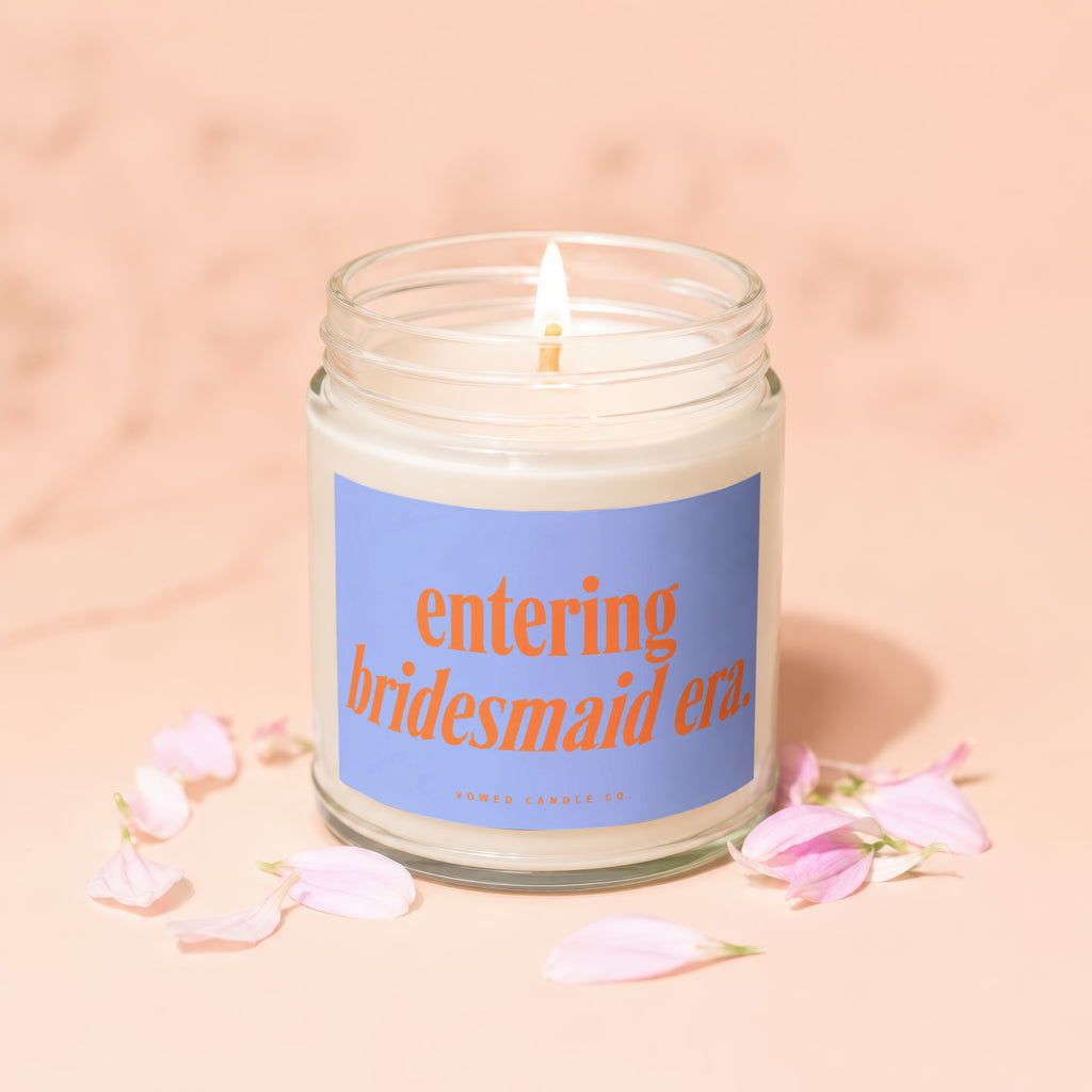 Glass jar candle with "entering bridesmaid era" text and pink petals on a peach background.