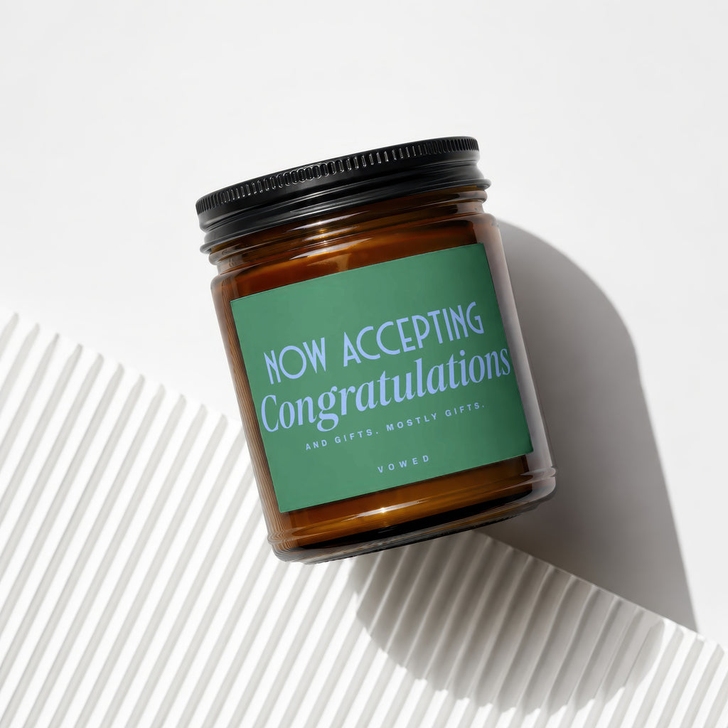 Jar candle with green label reading "NOW ACCEPTING Congratulations AND GIFTS. MOSTLY GIFTS. VOWED."