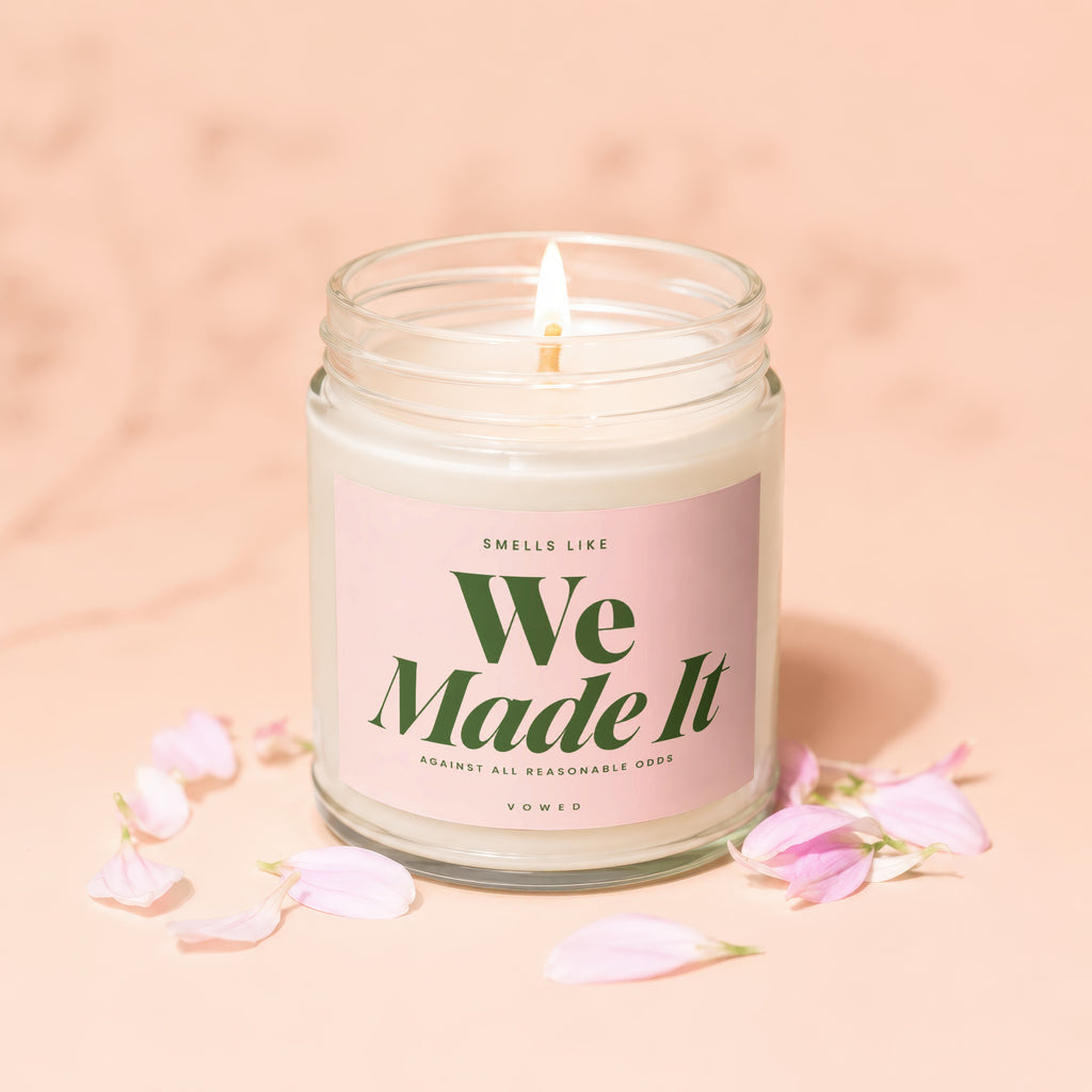 Lit candle with "We Made It" label and pink petals on a peach background.