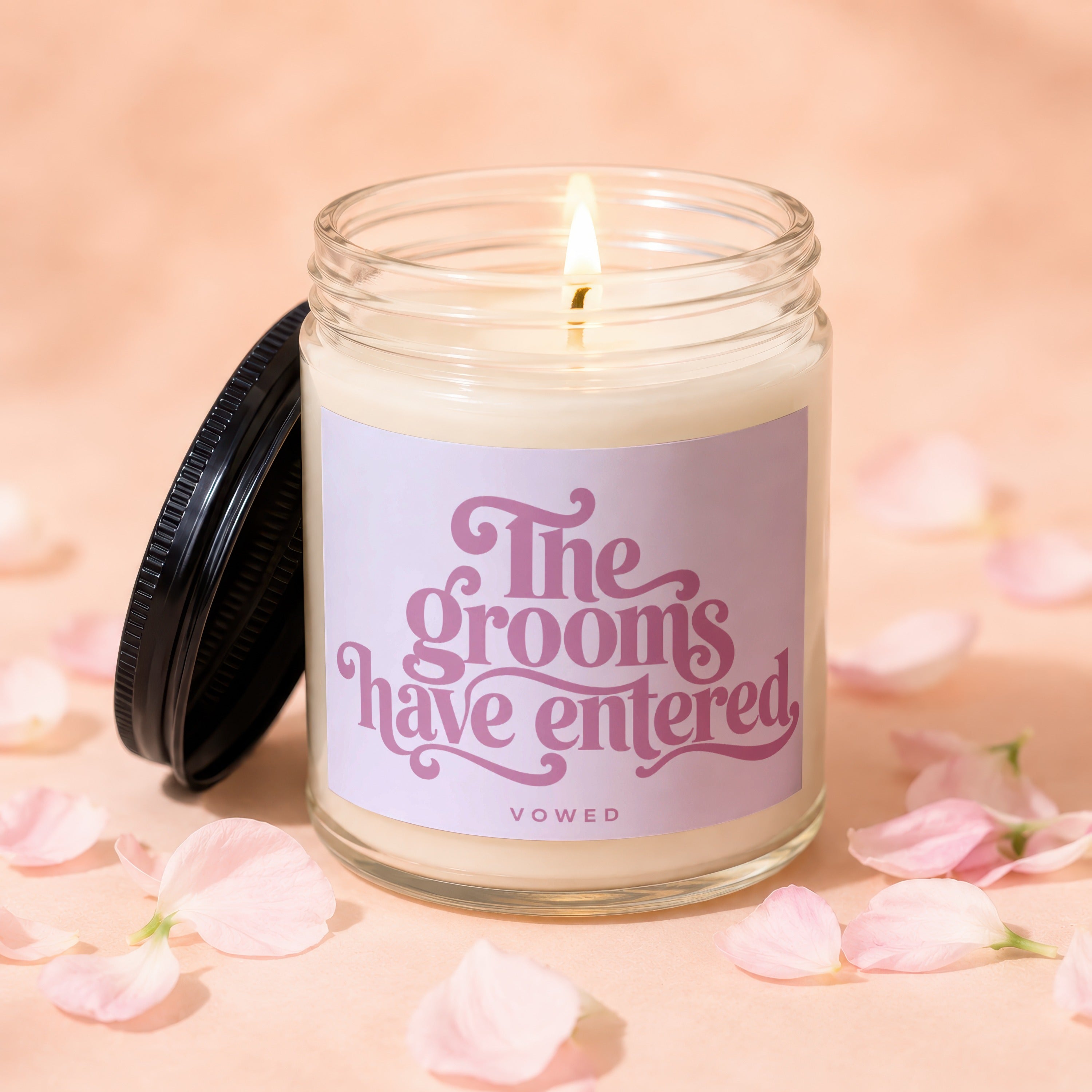Lit glass candle with pink label reading "The grooms have entered", on a peach background with petals.