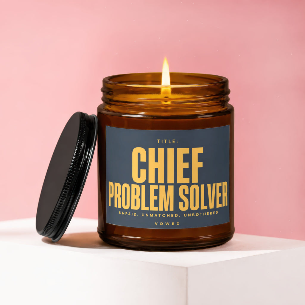 Brown glass candle with lit flame and dark lid, reading "Chief Problem Solver."