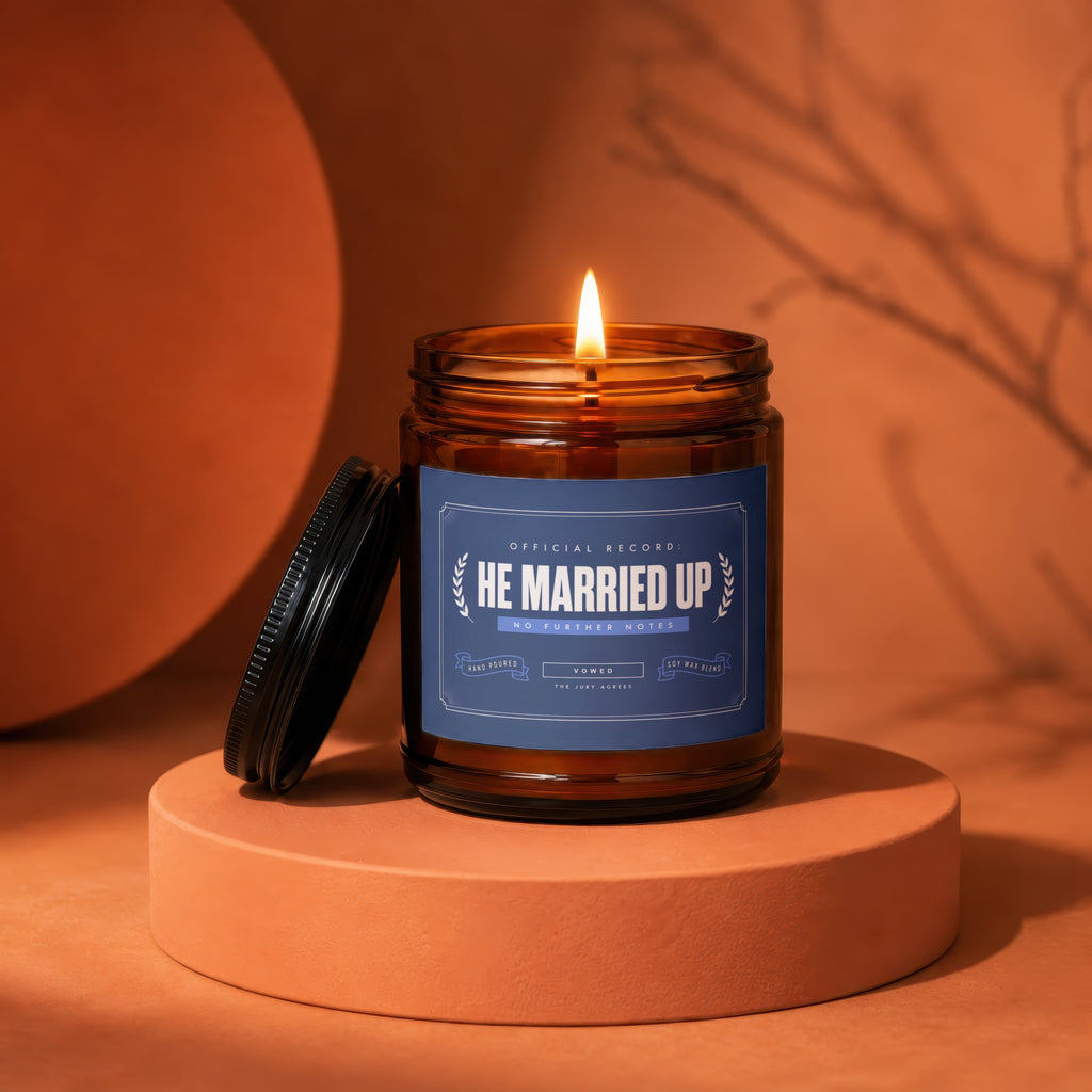 Amber glass candle with "He Married Up" label, lit flame, on an orange pedestal.