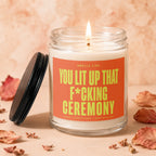 Glass candle with lit wick and orange label reading "Smells Like You Lit Up That F*cking Ceremony".