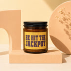 Amber jar candle with yellow label reading "HE HIT THE JACKPOT" and "VOWED CANDLE CO."