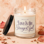 Candle with lit wick, lid off, reading "Love Is the Strongest Spell".