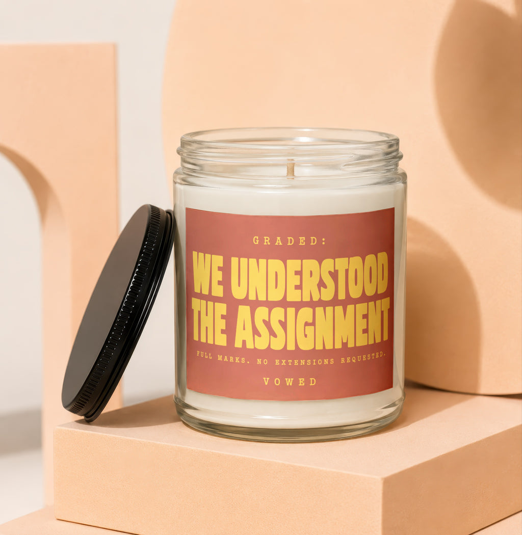 Glass jar candle with black lid, reads "GRADED: WE UNDERSTOOD THE ASSIGNMENT".