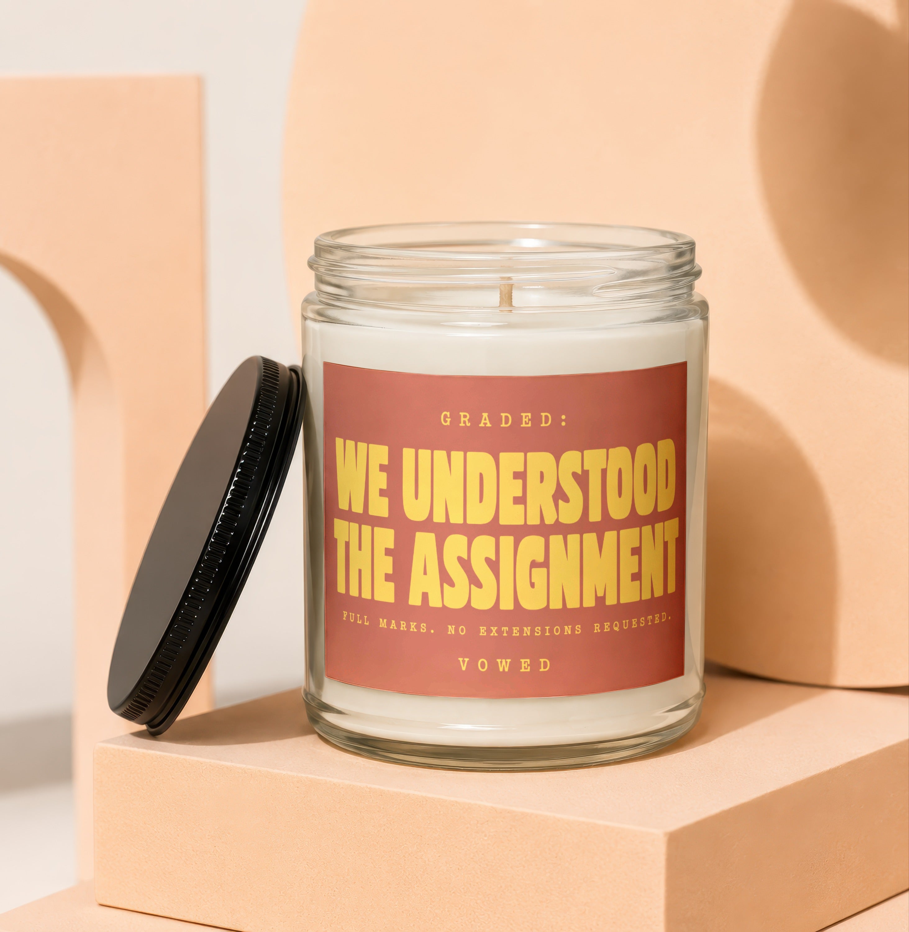 Glass jar candle with black lid, reads "GRADED: WE UNDERSTOOD THE ASSIGNMENT".