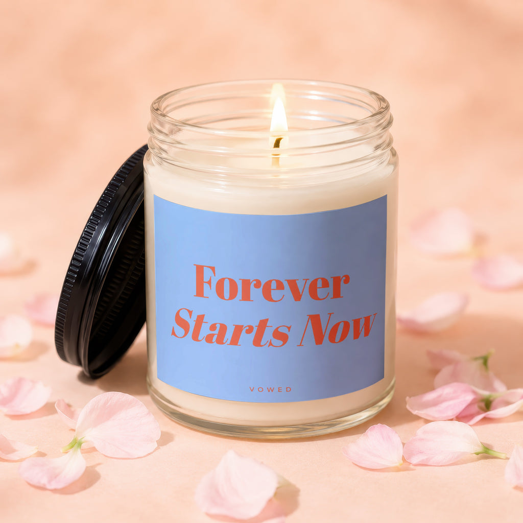 A lit candle in a glass jar with a blue label that reads "Forever Starts Now".