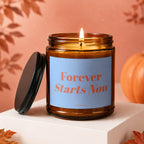 Amber jar candle with lit wick, blue label reads "Forever Starts Now".