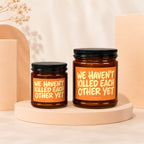 Two amber glass candles with orange labels reading "WE HAVEN'T KILLED EACH OTHER YET".