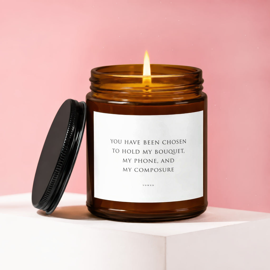 Amber jar candle with lit flame and lid off, labeled "You have been chosen to hold my bouquet, my phone, and my composure".