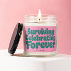 Glass jar candle with lit wick on pink background reads "Surviving Celebrating Forever Vowed".