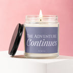 Candle with lit wick on a white surface, pink background, "The adventure Continues" text.