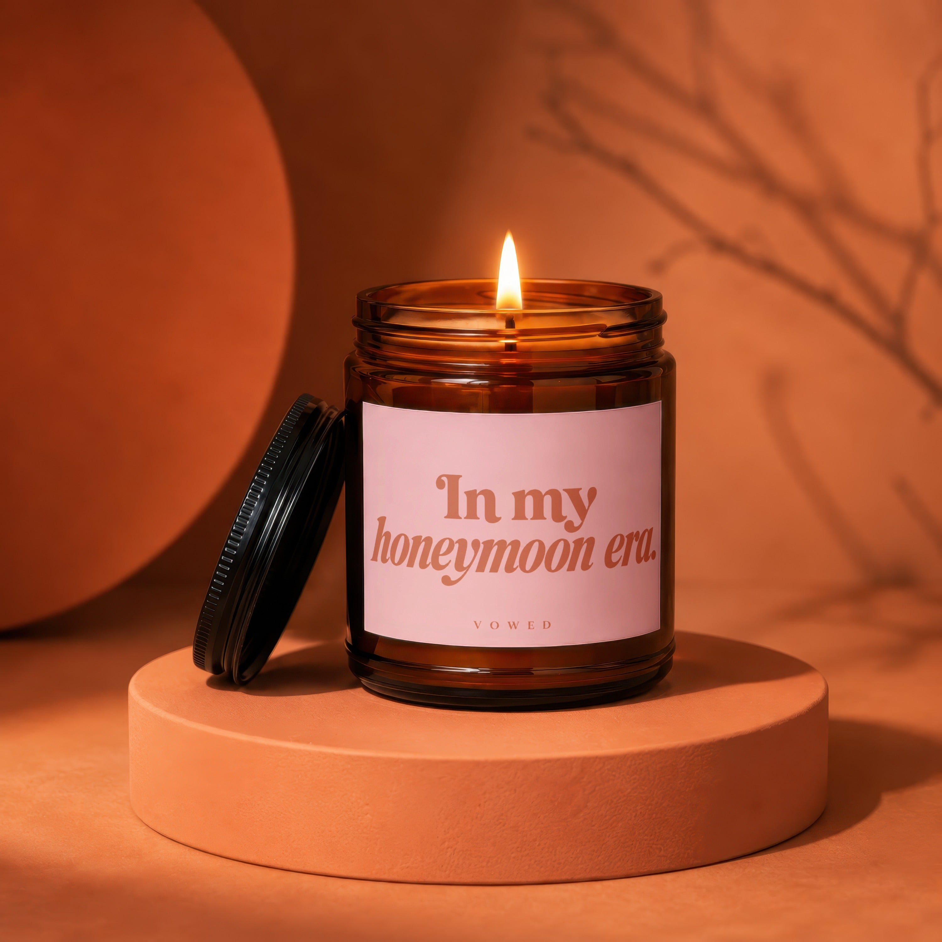 Brown glass candle with lit flame, pink label reading "In my honeymoon era. vo wed."