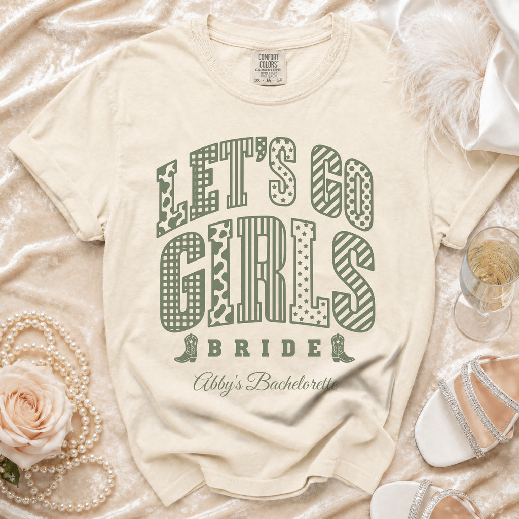 Cream-colored t-shirt with green text reading "LET'S GO GIRLS BRIDE Abby's Bachelorette" arranged among decorative elements.