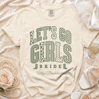 Cream-colored t-shirt with green text reading "LET'S GO GIRLS BRIDE Abby's Bachelorette" arranged among decorative elements.