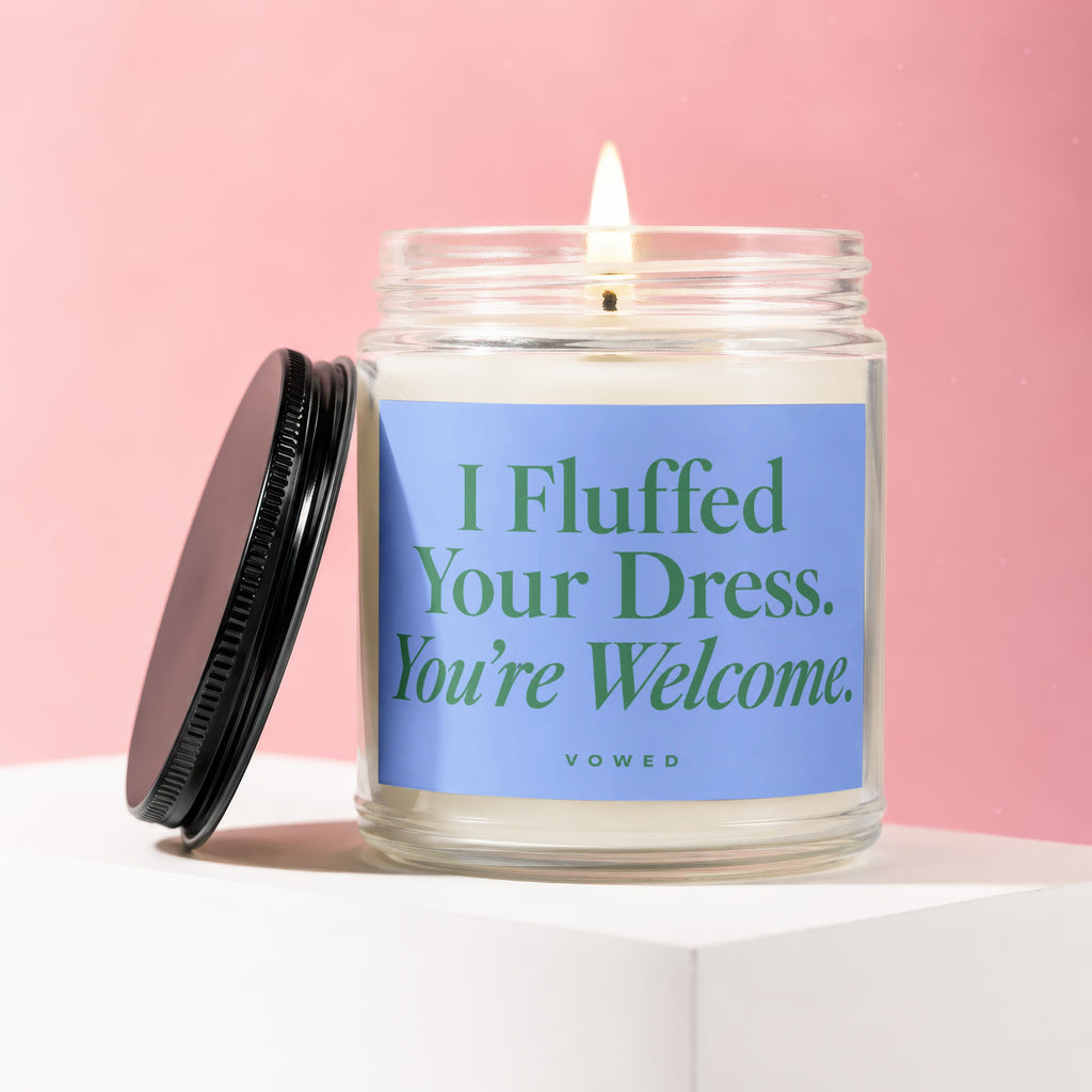 Jar candle with lid off, lit wick, reading "I Fluffed Your Dress. You're Welcome.", VOWED.