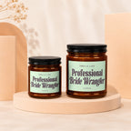 Two amber glass candles with black lids and mint green labels: "Smells Like Professional Bride Wrangler".